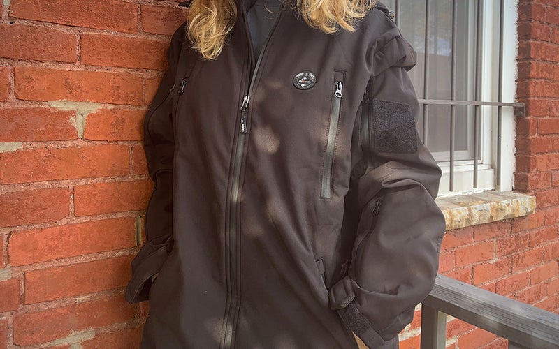 A person wearing an iHood Men's Heated Jacket with Detachable Hood outside while standing against a brick wall.
