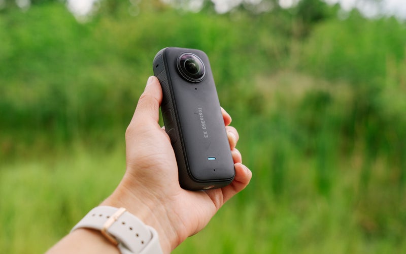 A hand holds the Insta360 X3 action camera in front of green grass.