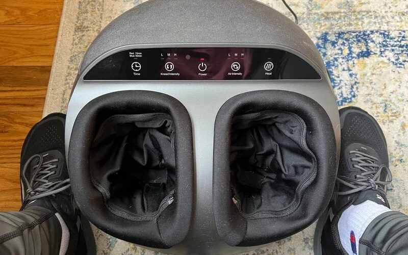 A gray Renpho Foot Massager with two pockets for feet on a carpet with two feet on either side.
