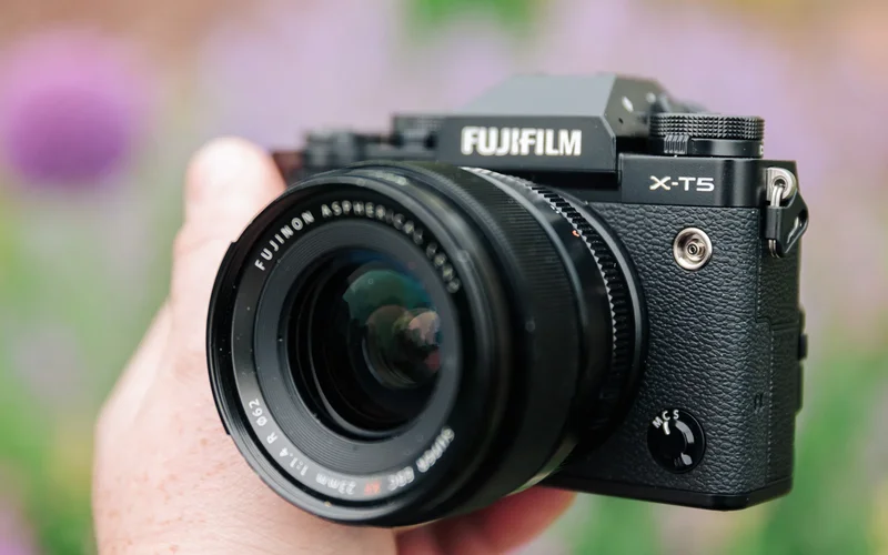 A hand holds the Fujifilm X-T5 mirrorless camera in front of a blurred background.
