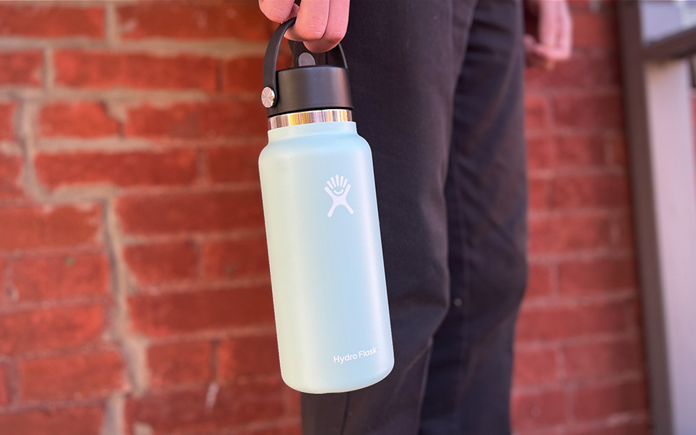 A person holding a blue Hydro Flask wide mouth bottle and standing in front of a brick wall.