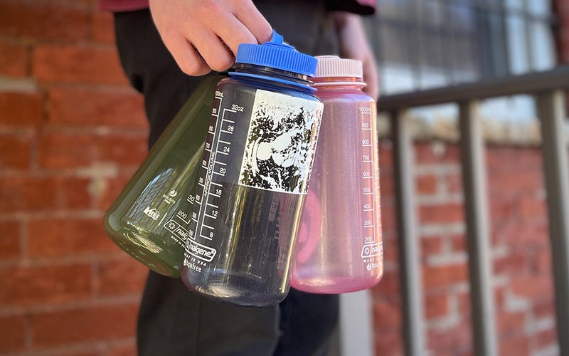 A person holding three Nalgene water bottles and standing against a brick wall.