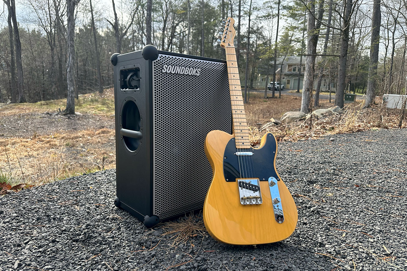 Black Soundboks 4 outdoors speaker with an orange Fender guitar on a gravel patio
