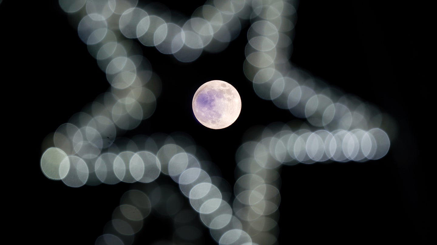 The last full moon of 2022 rises through a clear sky, past a string of festive lights. The moon is positioned at the center of a star made up of white Christmas lights.