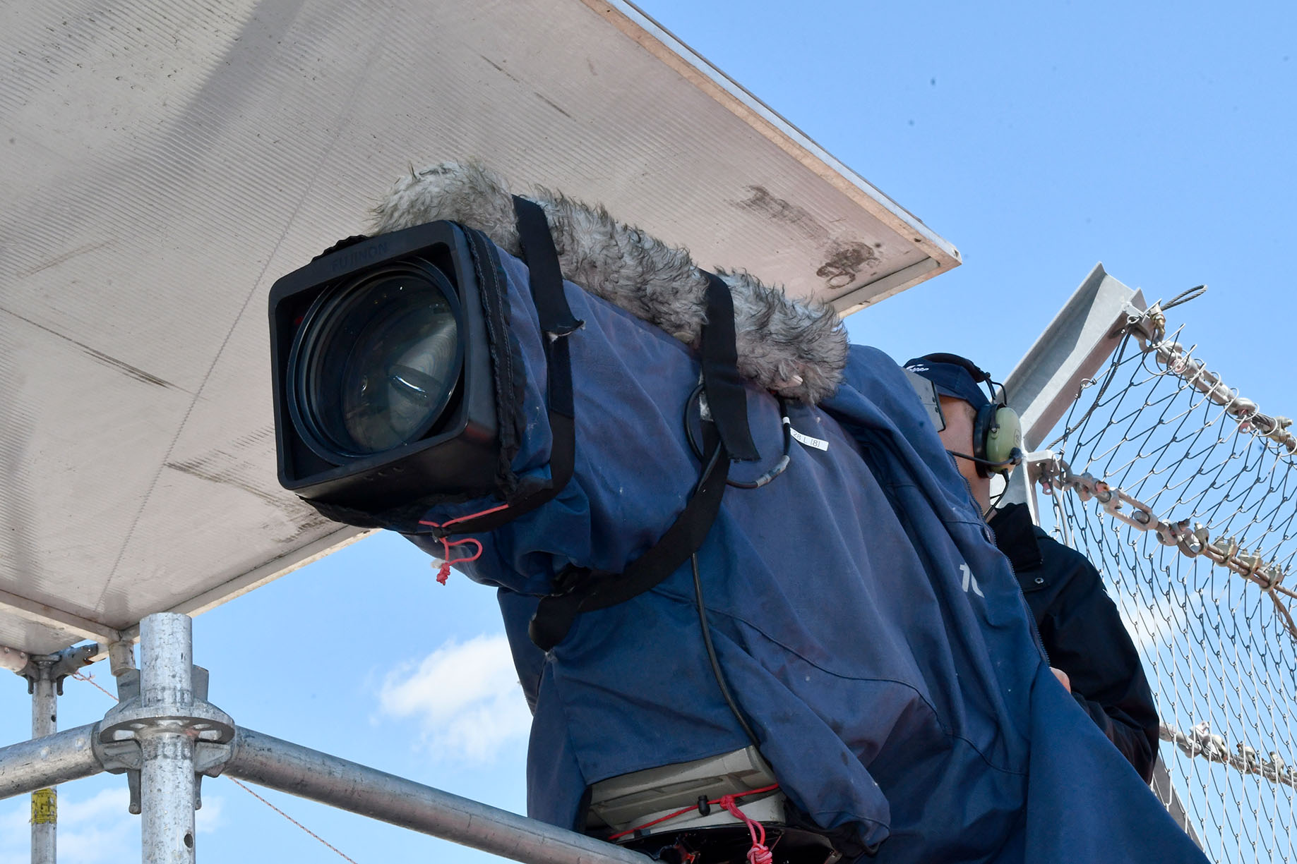 A trackside broadcast camera on a riser in a blue weather-resistant wrap topped by an Audio Technica shotgun mic in a windshield