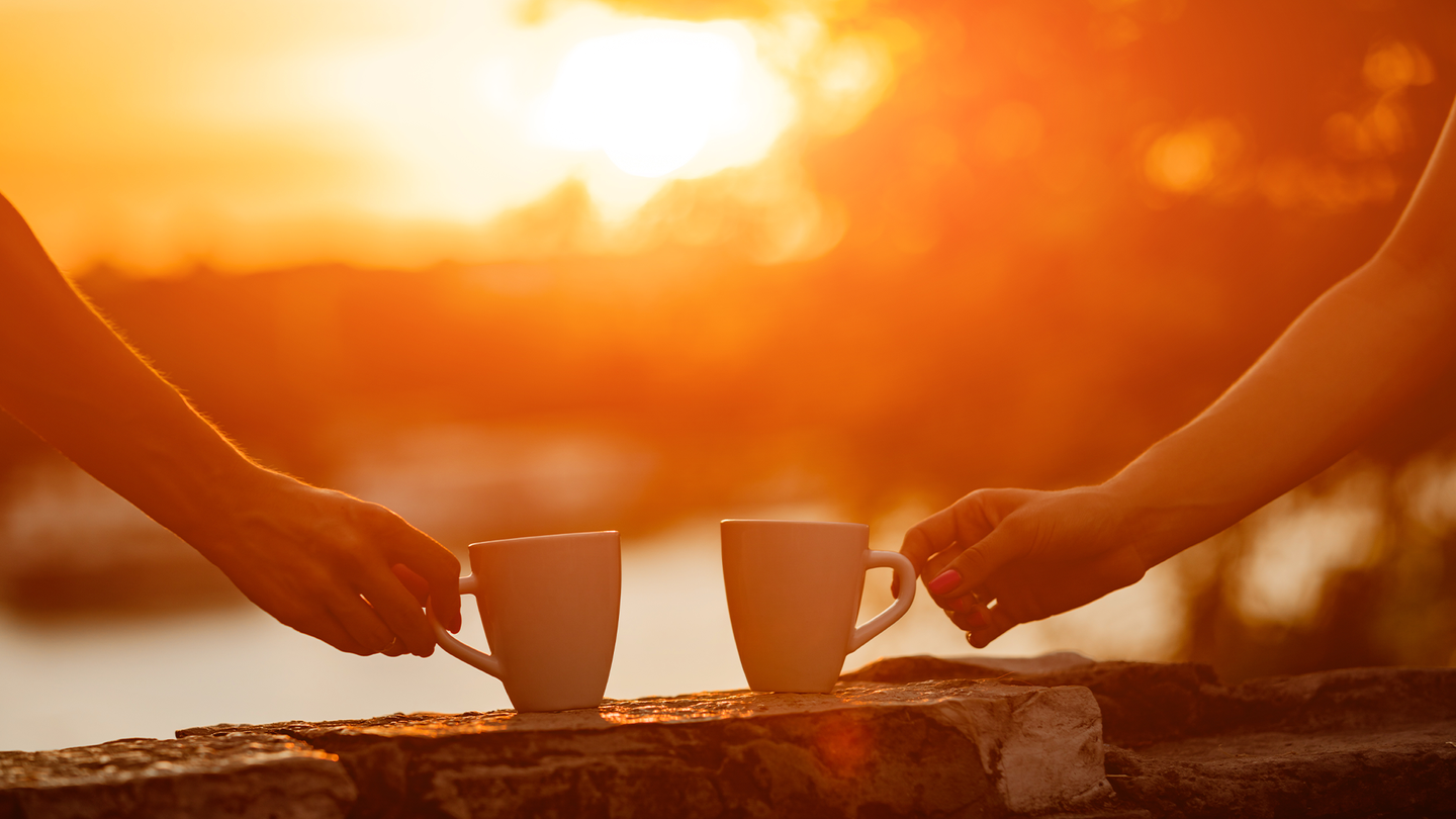 Two people hold coffee cups while looking at a a sunrise.
