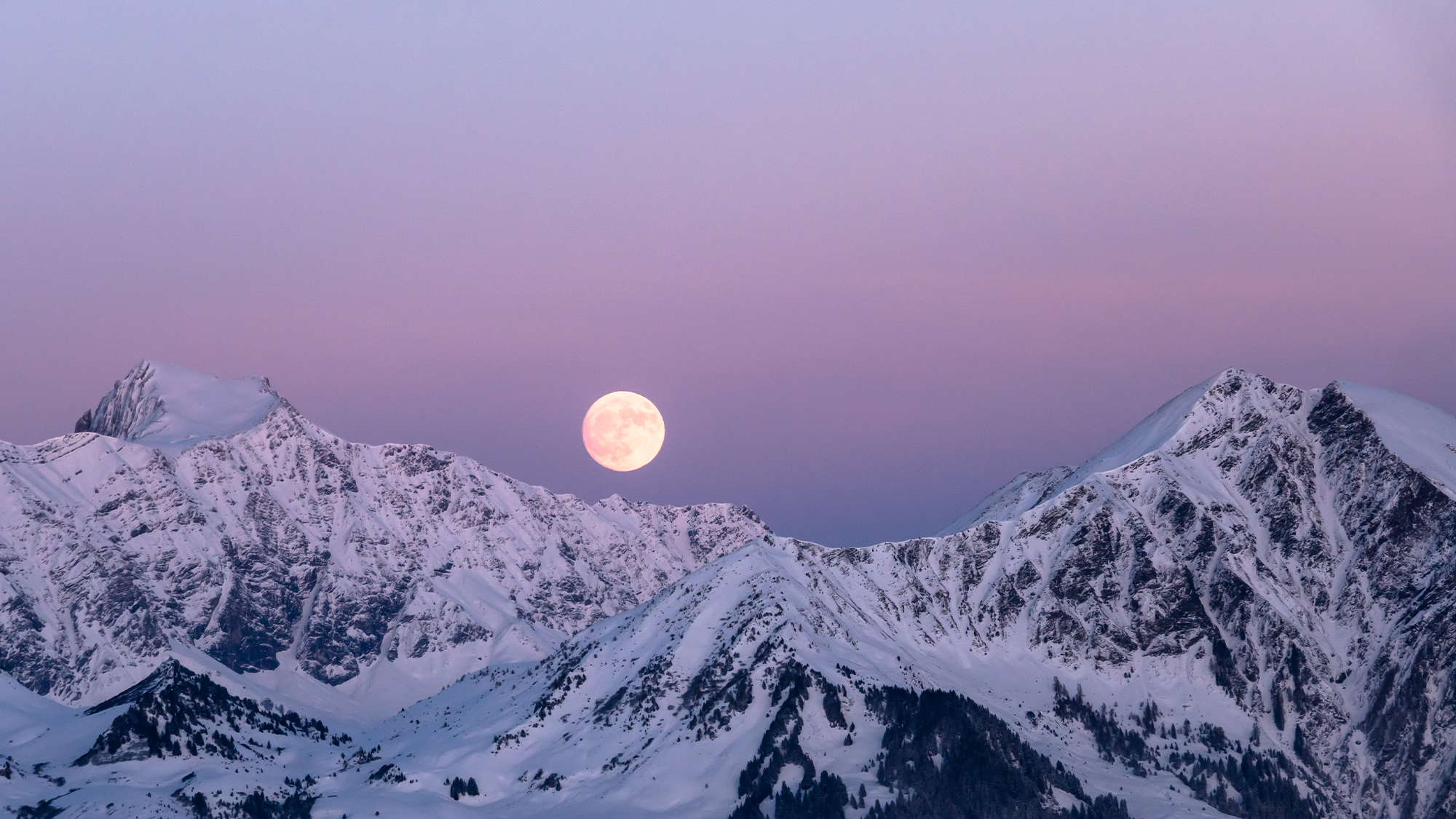 A full moon rises n a purple sky over snowy mountain peaks.