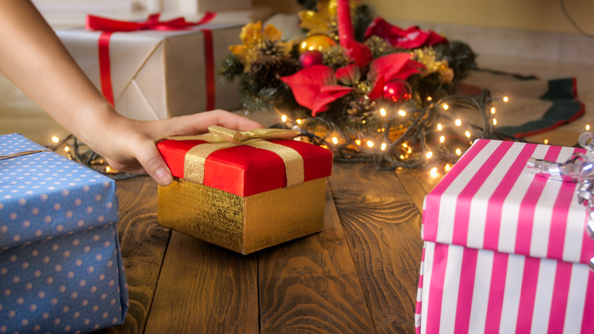 A hand reaches for a present wrapped in red and gold wrapping paper underneath a Christmas tree.