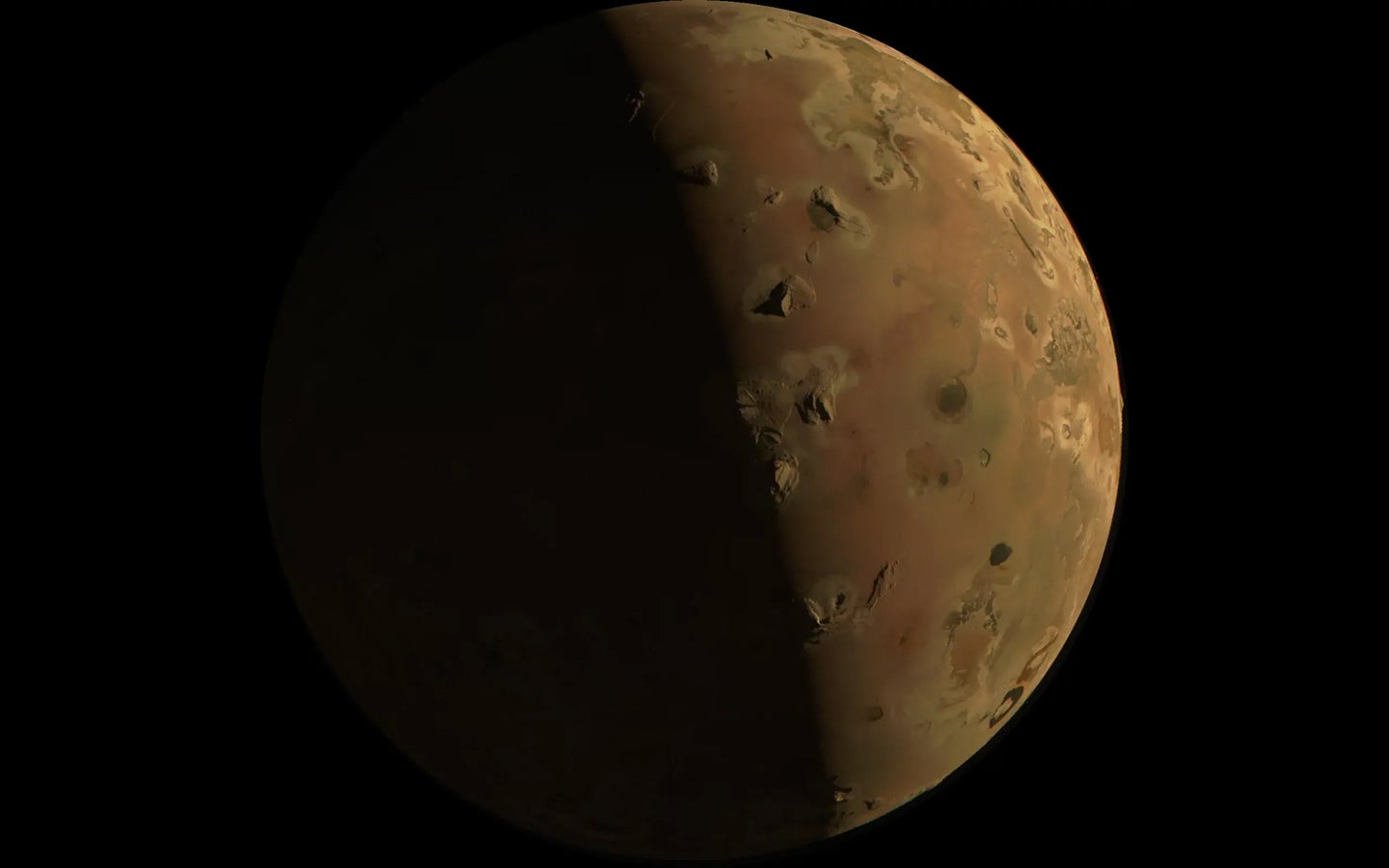 A reddish moon on a black background, dotted with several gray marks that are volcanoes. Jupiter's moon Io is one of Jupiter’s several moons. This image was taken on December 30, 2023 during the Juno spacecraft’s flyby of this volcanic Jupiterian moon.