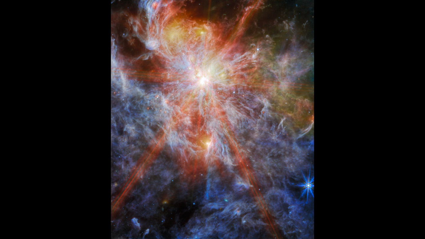 A bright young star within a colorful nebula. The star is identifiable as the brightest spot in the image, surrounded by six large spokes of light that cross the image. A number of other bright spots can also be seen in the clouds, which are shown in great detail as layers of colorful wisps.