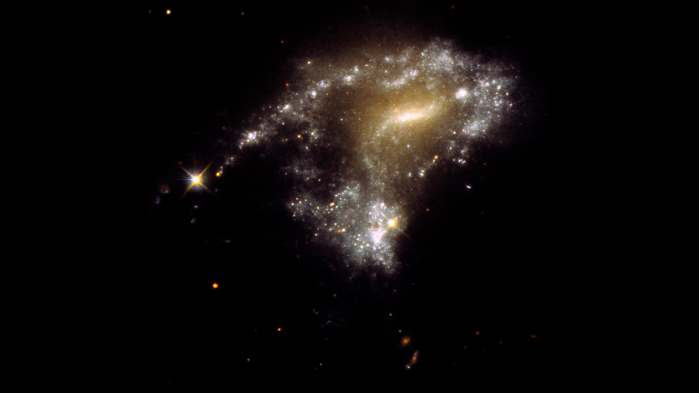 Galaxy AM 1054-325 has been distorted into an S-shape from a normal pancake-like spiral shape by the gravitational pull of a neighboring galaxy, seen in this NASA Hubble Space Telescope image. A consequence of this is that newborn clusters of stars form along a stretched-out tidal tail for thousands of light-years, resembling a string of pearls. They form when knots of gas gravitationally collapse to create about one million newborn stars per cluster.