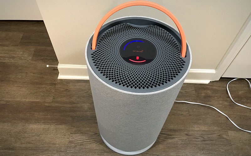 Grey round Oransi Mod air purifier with an orange handle on wood floor in front of white walls