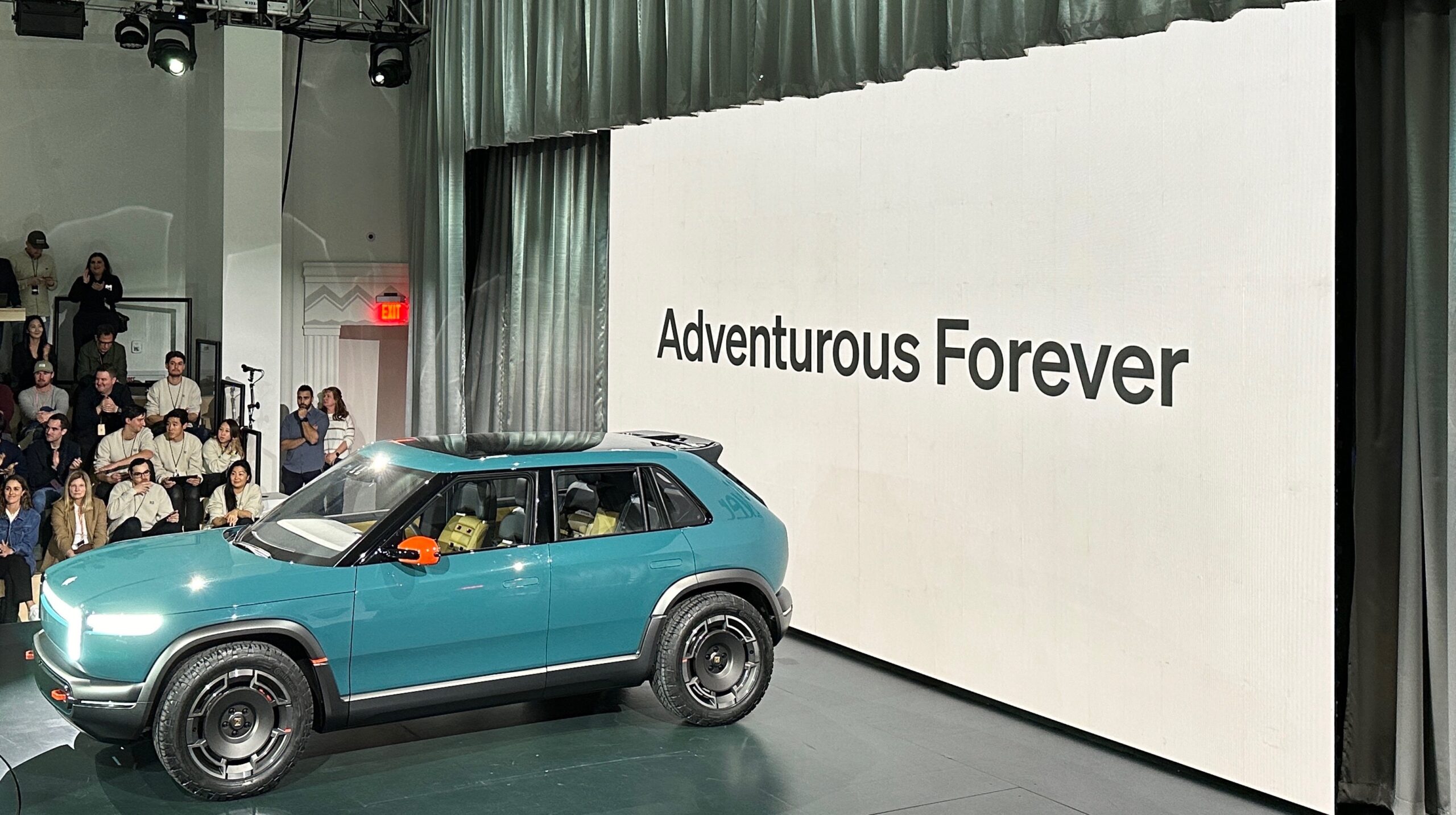 a green-blue hatchback car on a stage in front of the words "Adventurous Forever"