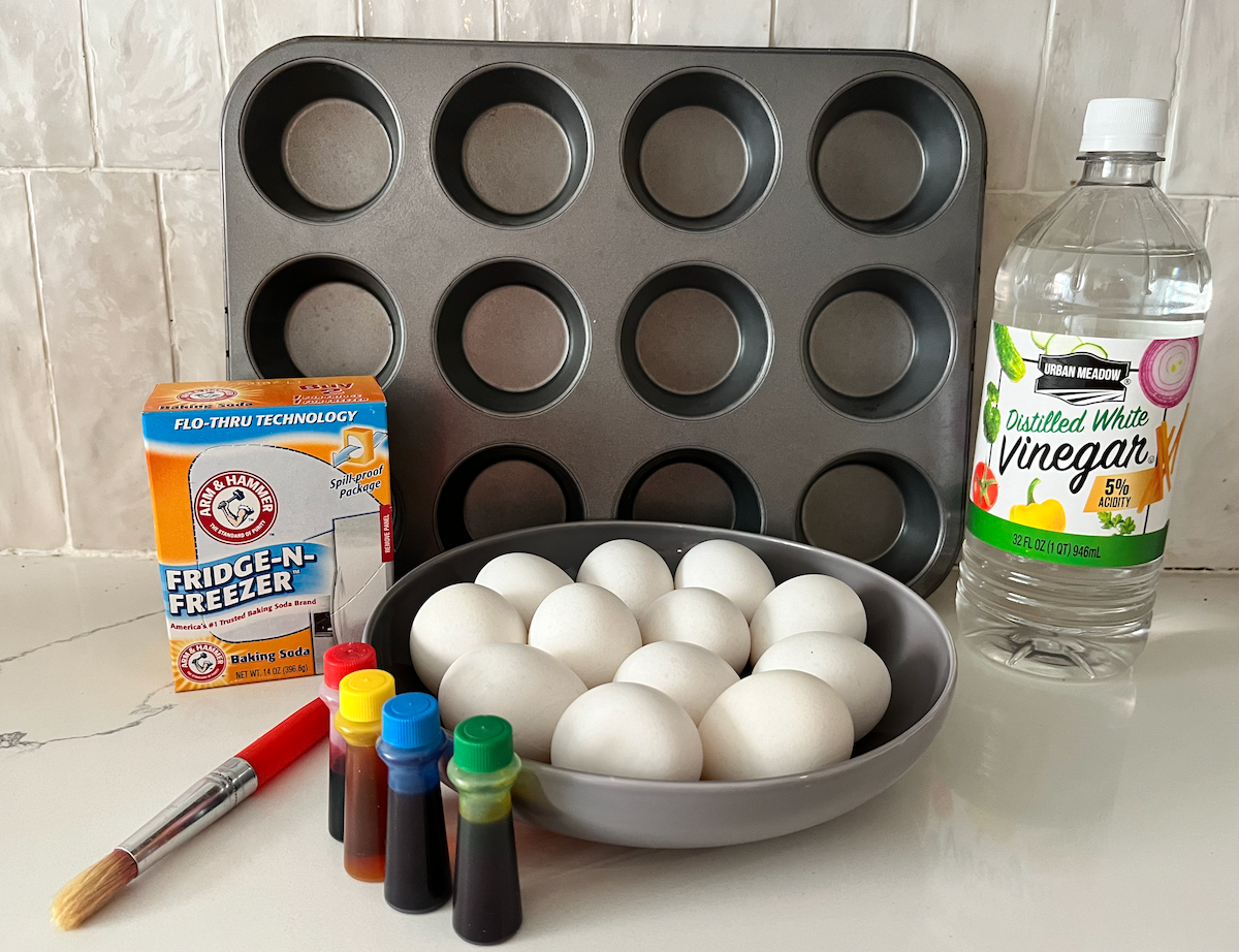 muffin tin, baking soda, vinegar, a dozen eggs, paint brush, and food coloring on a white countertop