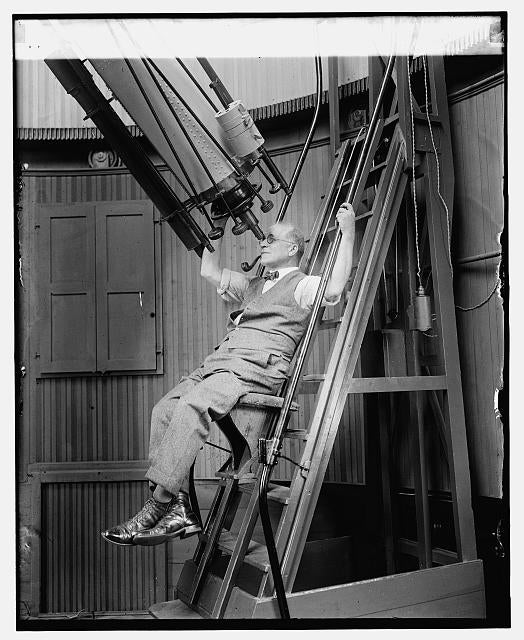 a man with a pipe and bowtie sits on a ladder looking through a large telescope