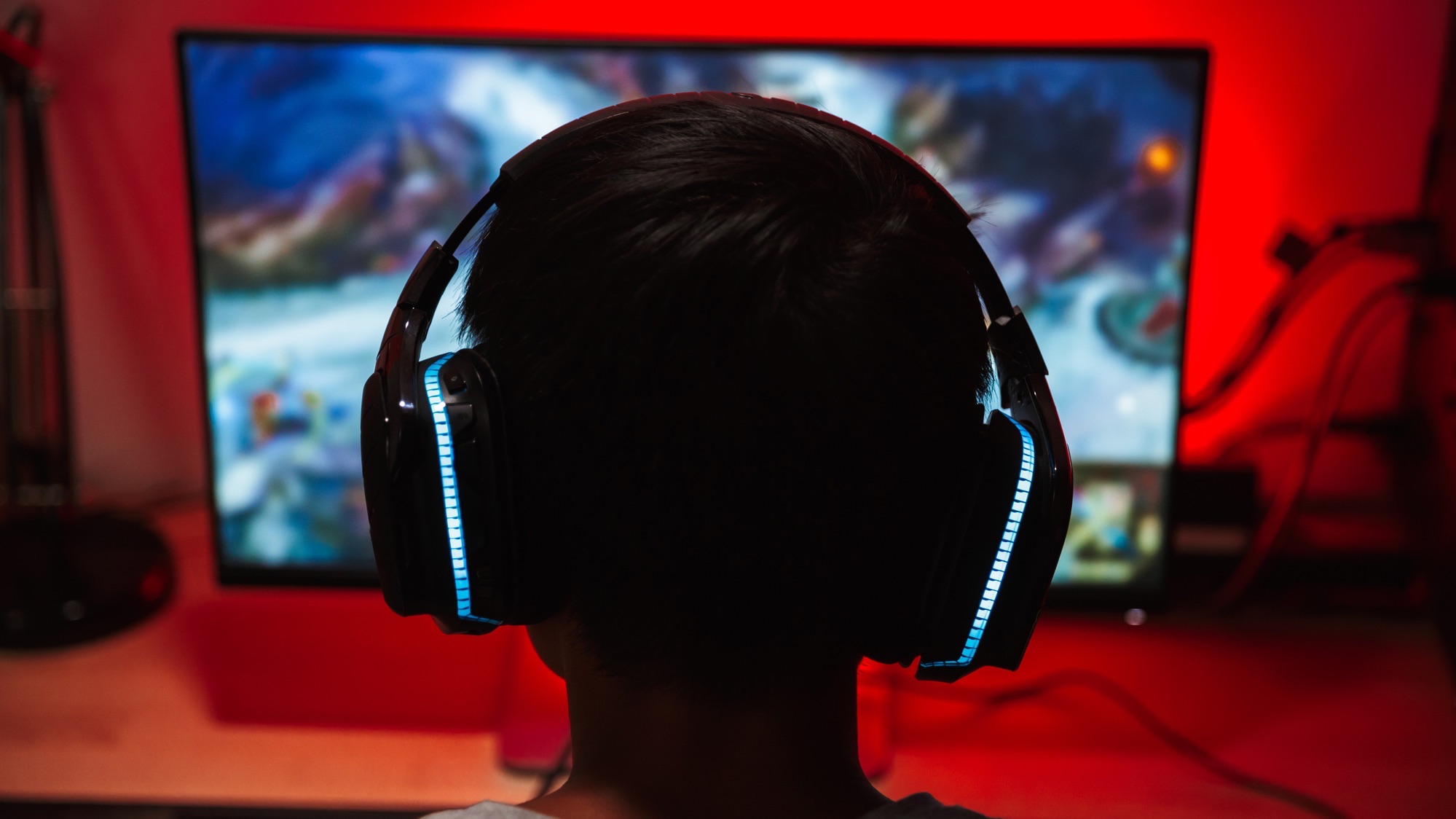 Portrait from back of young gamer guy looking at screen, and playing video games on computer in dark room wearing headphones