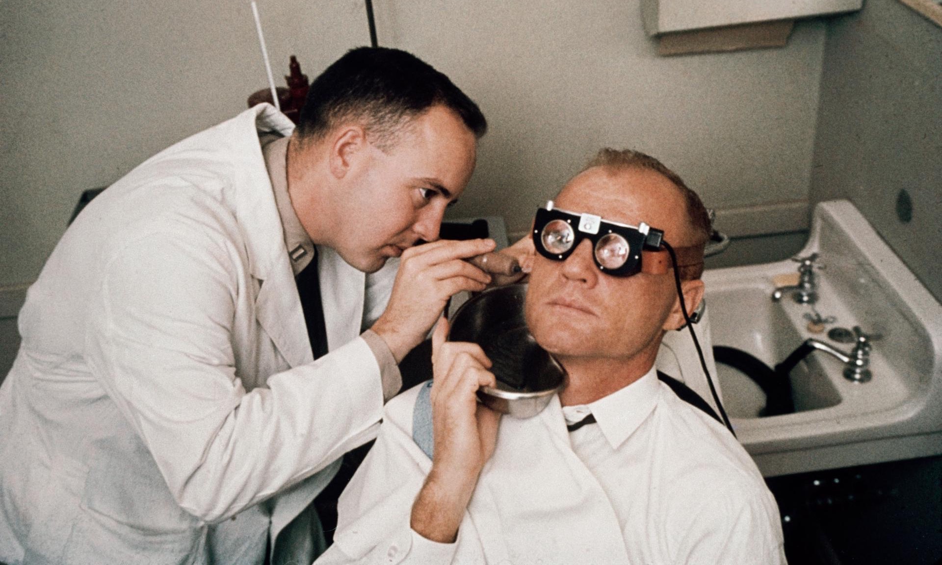 a man looks into the ear of a man with thick glasses on