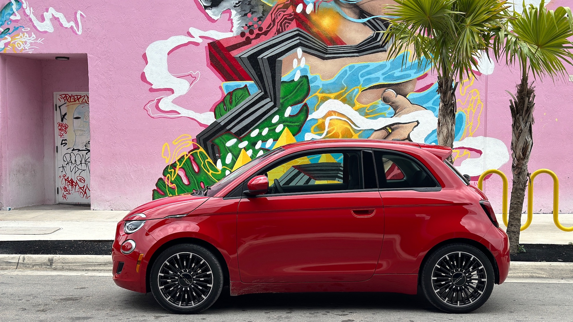 small red car sits in front of a pink wall covered in art