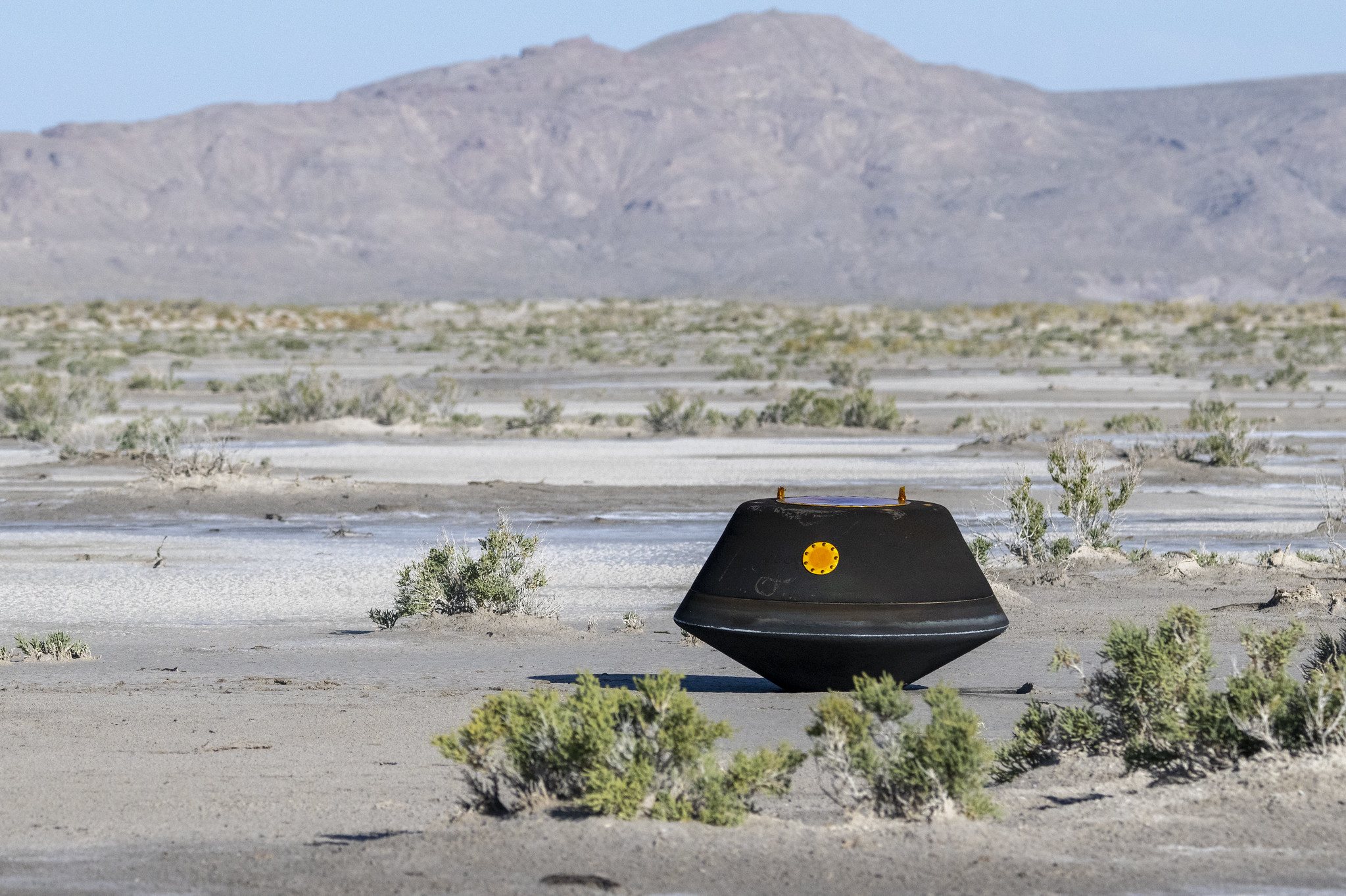 a black capsule sits on desert sand near shrub brush