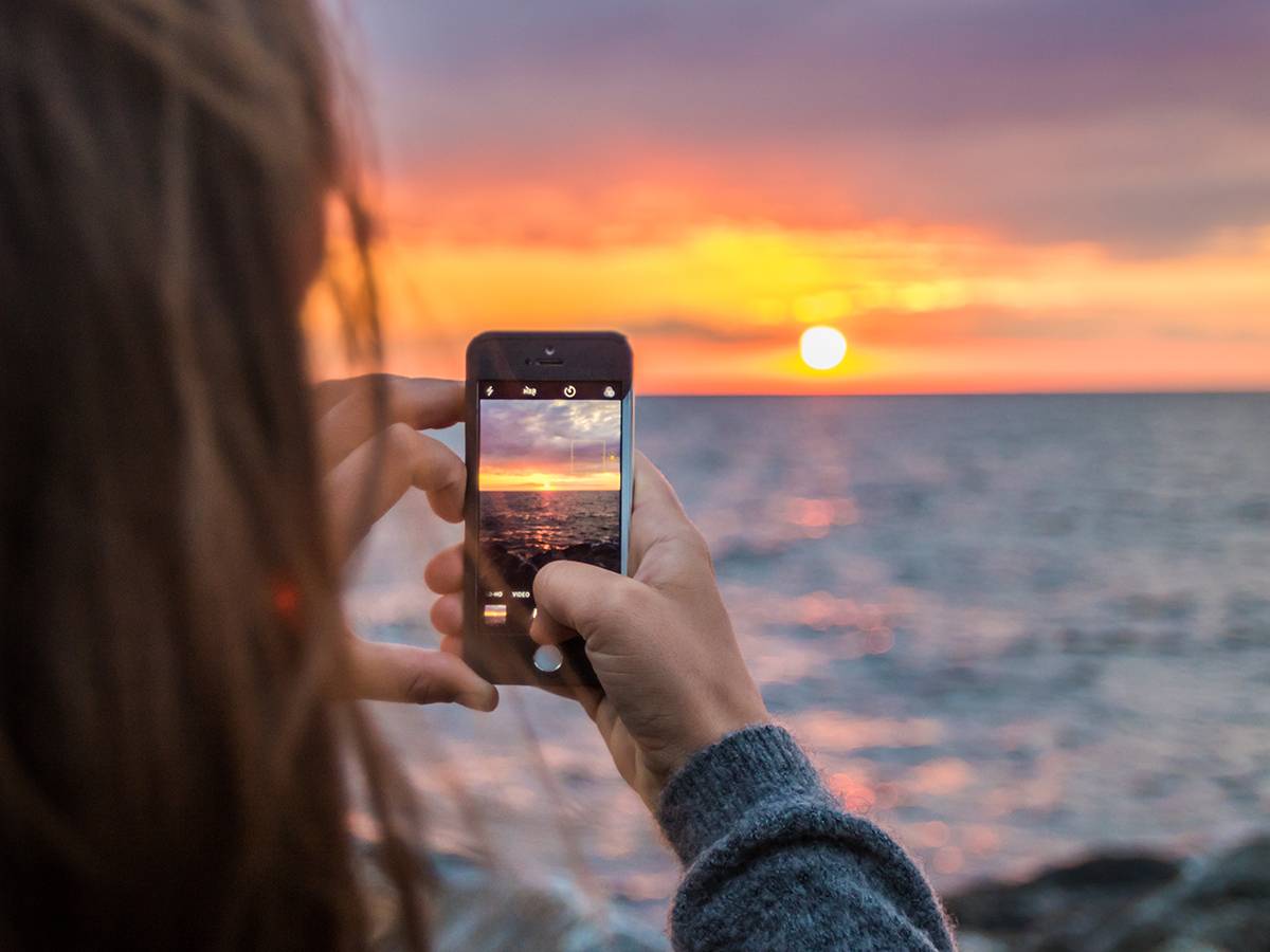A woman standing by the ocean, using her phone to take a photo of the sunset over the water.