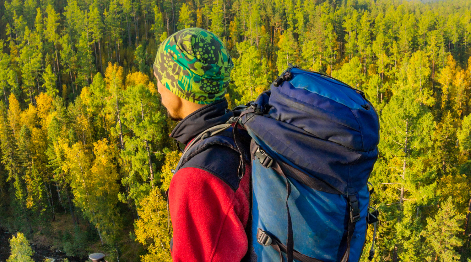 Man with backpack and trekking pole in bandana standing on a rock at dawn above a background autumn forest.