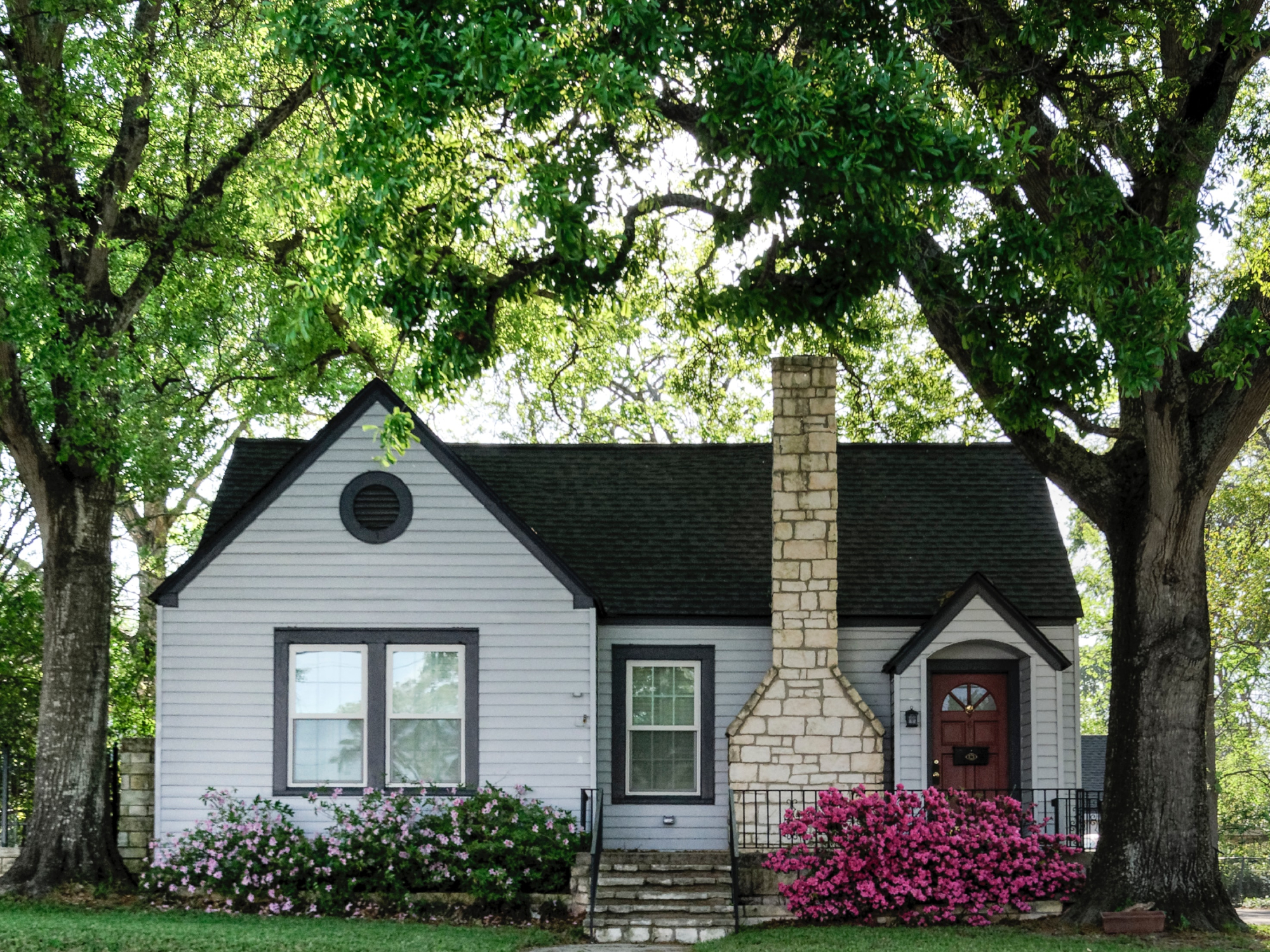 A small, white one-story house with a blooming garden, under some large green trees.