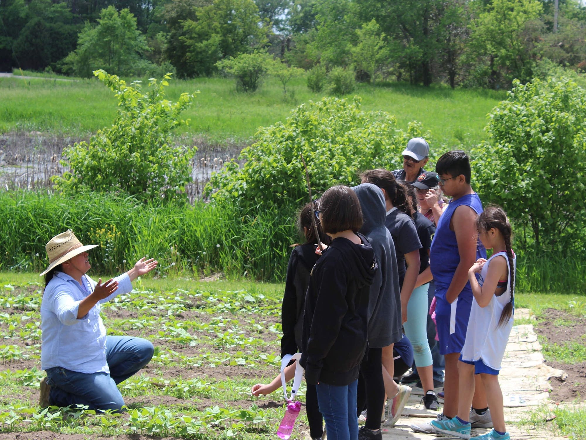 Indigenous farmer in a sun hat kneeling and speaking to a group of children in a green field in summer