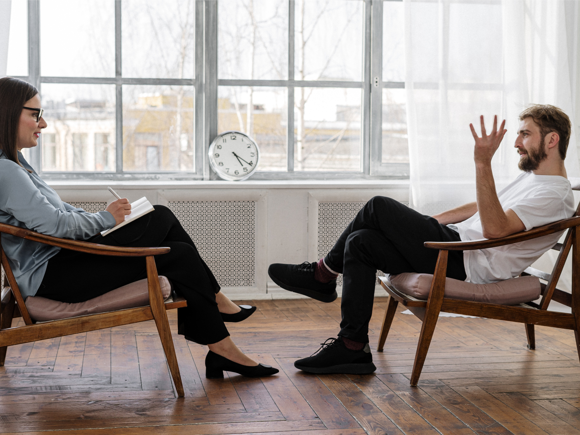 A man in a white t-shirt and black pants sitting in a chair talking to a therapist while they both sit in front of large windows with white curtains.
