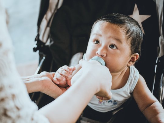 A baby in a stroller drinking formula from a bottle held by a woman.