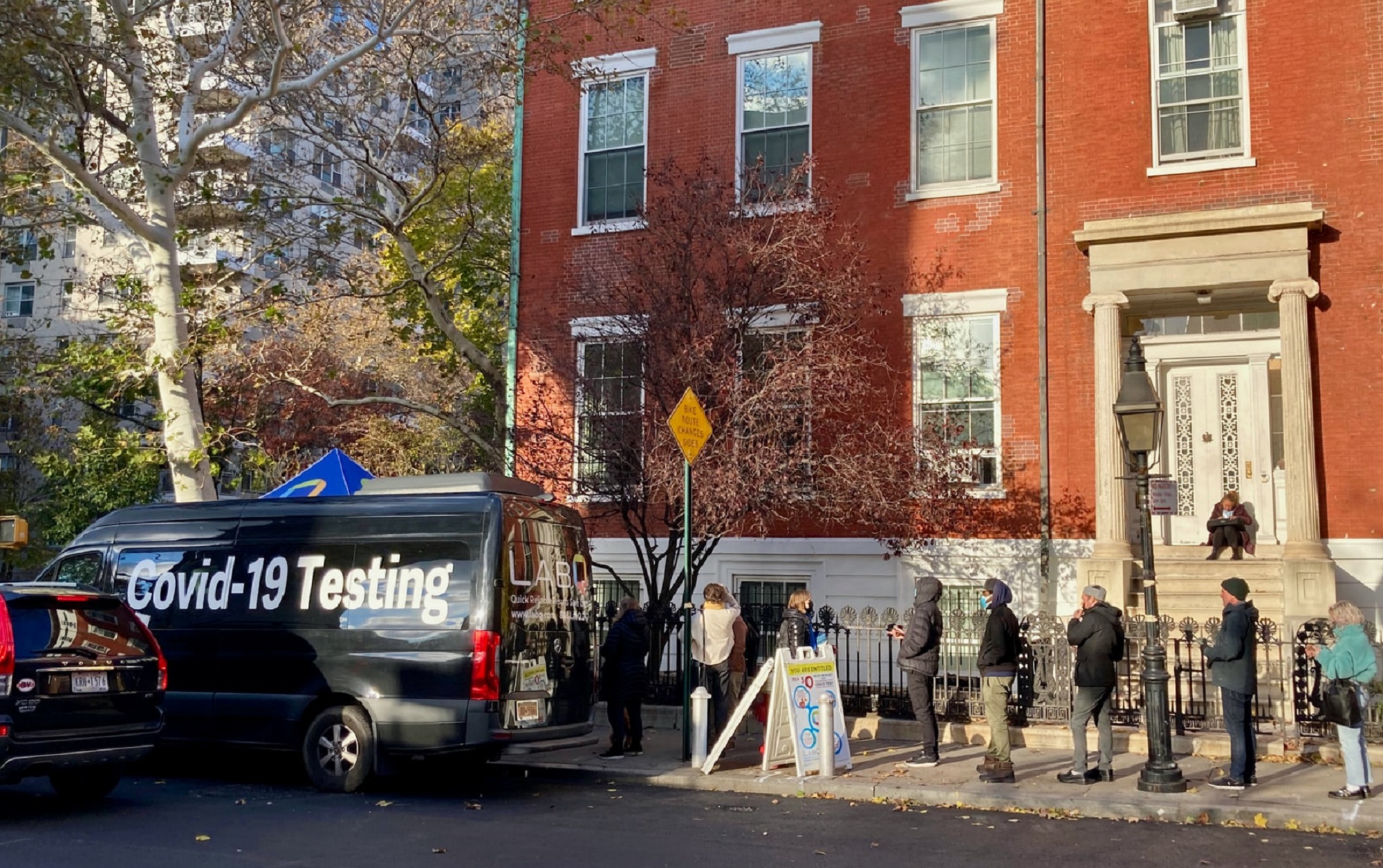 People in masks waiting on a New York City street in daytime for free COVID-19 testing at a blue truck