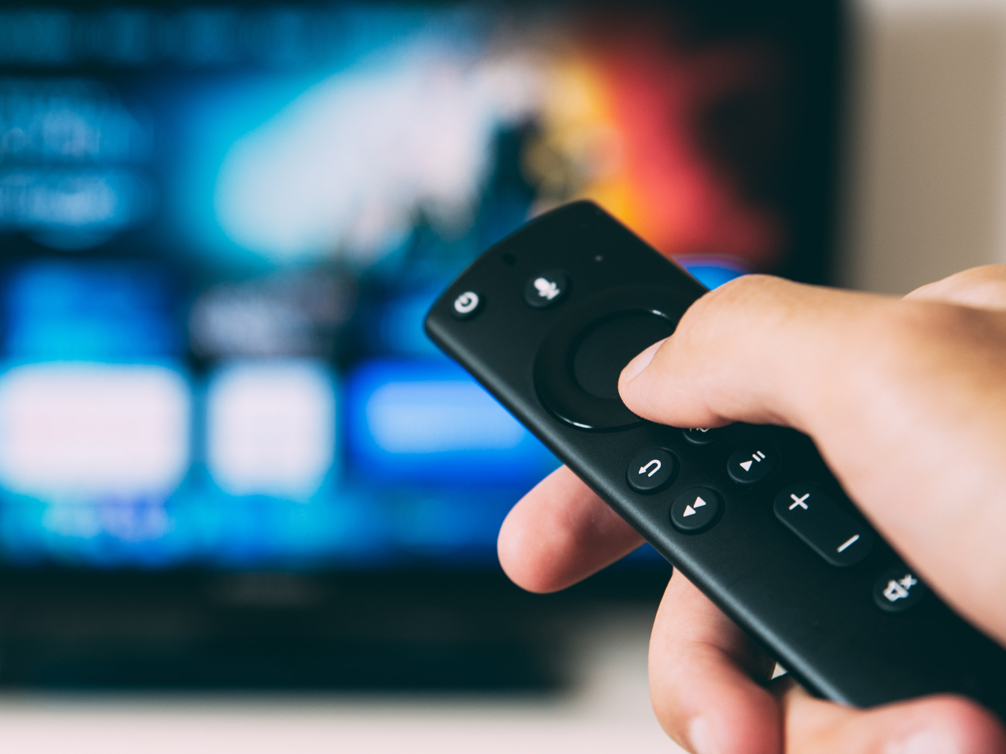 A close-up of a hand holding a TV remote with an out-of-focus TV screen in the background.