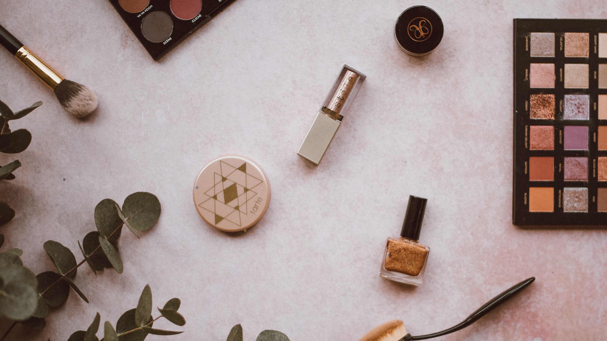 Various types of makeup on a white surface, with some leaves nearby.