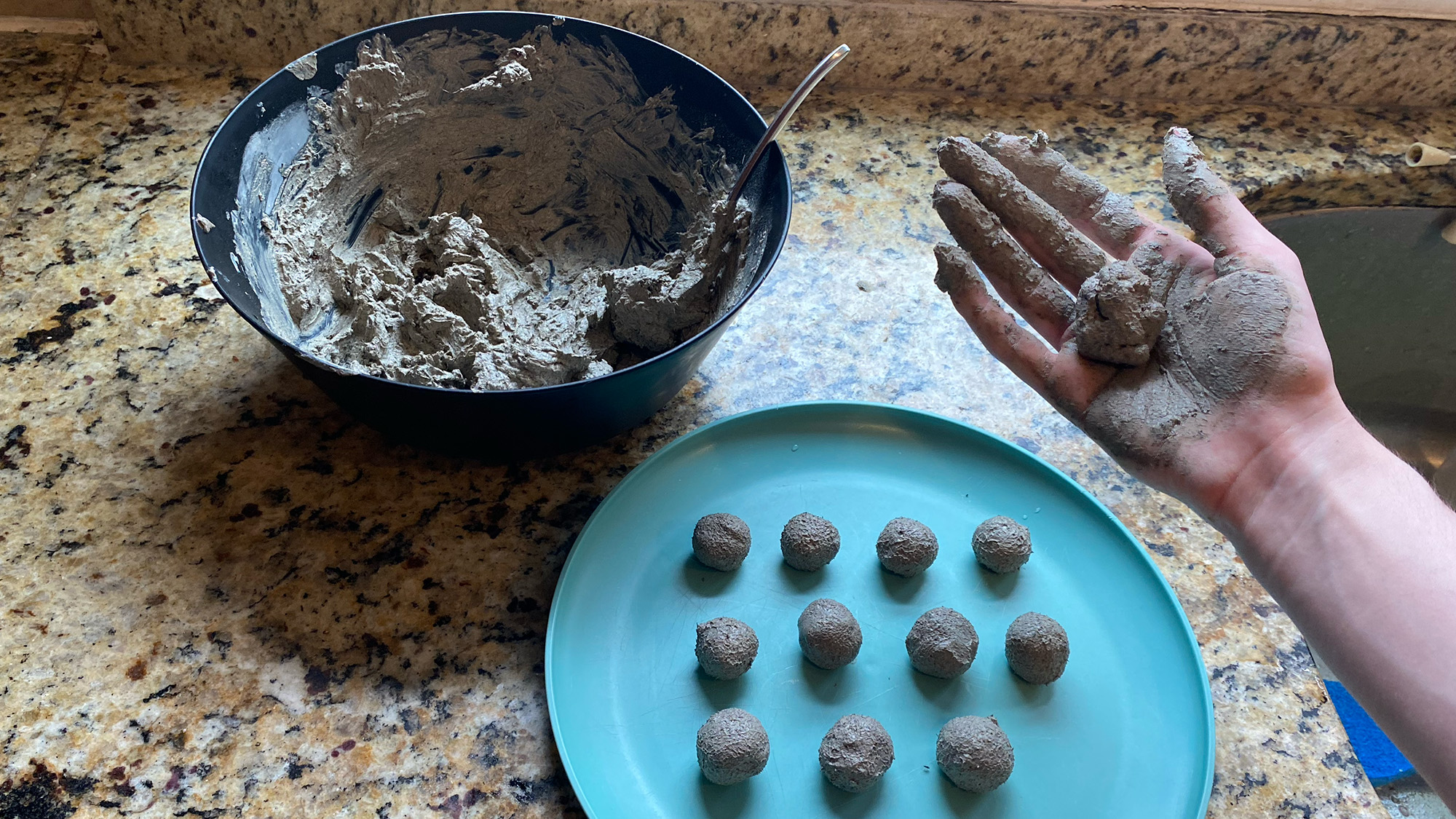 Kitchen counter with a bowl of seed bomb mix, a plate with drying bombs, and a hand holding an unfinished bomb.