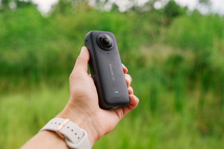 A hand holds the Insta360 X3 action camera in front of green grass.