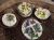 various easter cheese and butters are on a table. a cross made from butter is laid on a bed of greens with purple flowers. a round cheese is sprinkled with greens. a dish with wet cheese is decorated with yellow and white flowres. a scallop bowl of butter balls is decorated with greens.