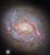 A close-up of a spiral galaxy, seen face-on. Its center glows brightly. From the sides of the galaxy’s core emerge spiral arms which wind through the round disc of the galaxy, filled with shining pink spots where stars are forming and more dark-red dust. Some faint stars can be seen around the galaxy, as well as a particularly bright star in the lower left of the image.