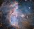 A star cluster within a nebula. The background is filled with thin, pale blue clouds. Parts are thicker and pinker in colour. The cluster is made up of bright blue stars that illuminate the nebula around them. Large arcs of dense dust curve around, before and behind the clustered stars, pressed together by the stars’ radiation. Behind the clouds of the nebula can be seen large numbers of orange stars.