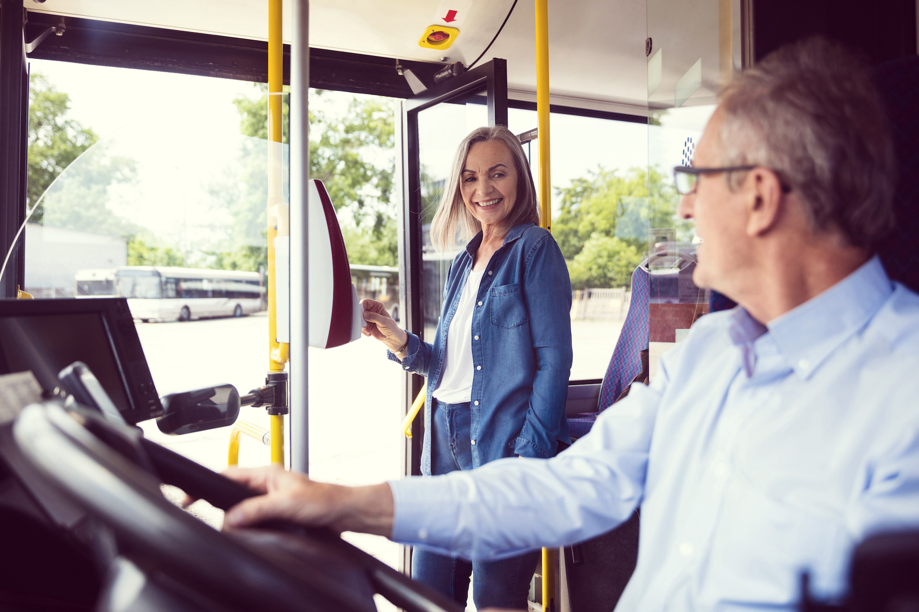 Passenger making contactless payment in public transport. Senior male driver is driving bus. Elderly woman is standing at entrance.