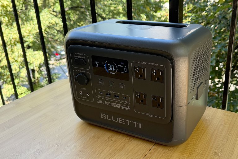 A grey Bluetti Elite 100 portable power station sitting on a wooden balcony table with a black railing and green leaves behind it