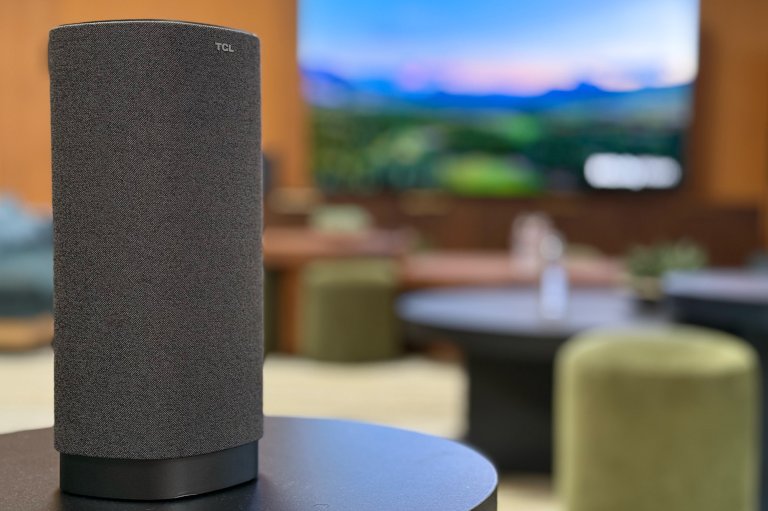 A grey TCL Z100 speaker on a side table, with colorful couches and ottomans in the background