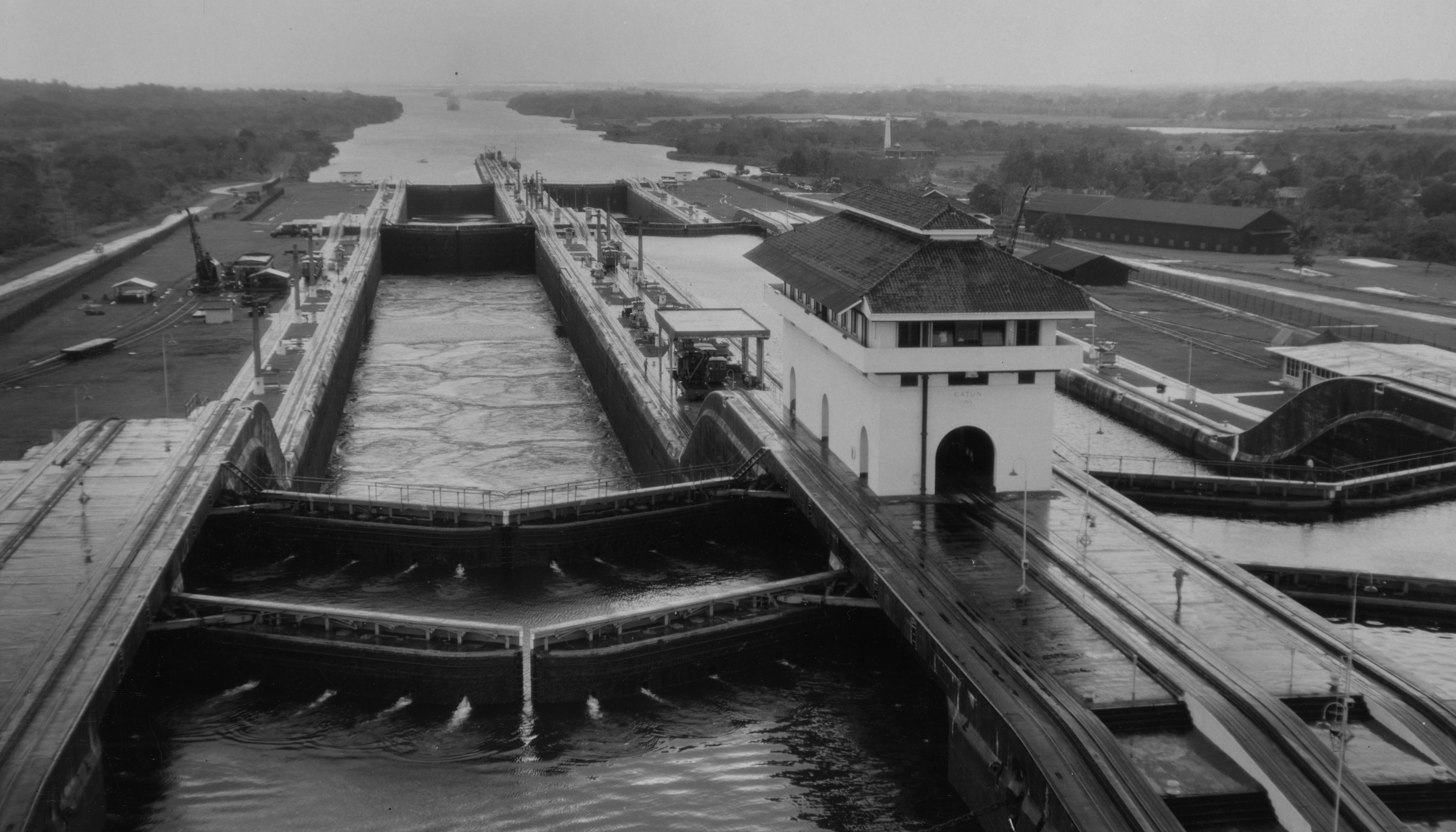 A black and white, elevated view of the Gatun Locks on the Panama Canal. The massive concrete lock chambers are filled with water, and a large white control building with a dark roof stands on the right side of the locks. Long, narrow bridges cross over the lock gates. In the distance, a large body of water, likely a lake or another section of the canal, stretches towards the horizon, bordered by forested land. Various structures and equipment are visible along the edges of the locks.