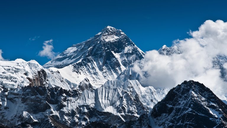 A dramatic, high-contrast shot of the snow-capped summit of Mount Everest, with jagged ridges and a glacier visible below. The peak is set against a deep blue sky and partially surrounded by bright white clouds.