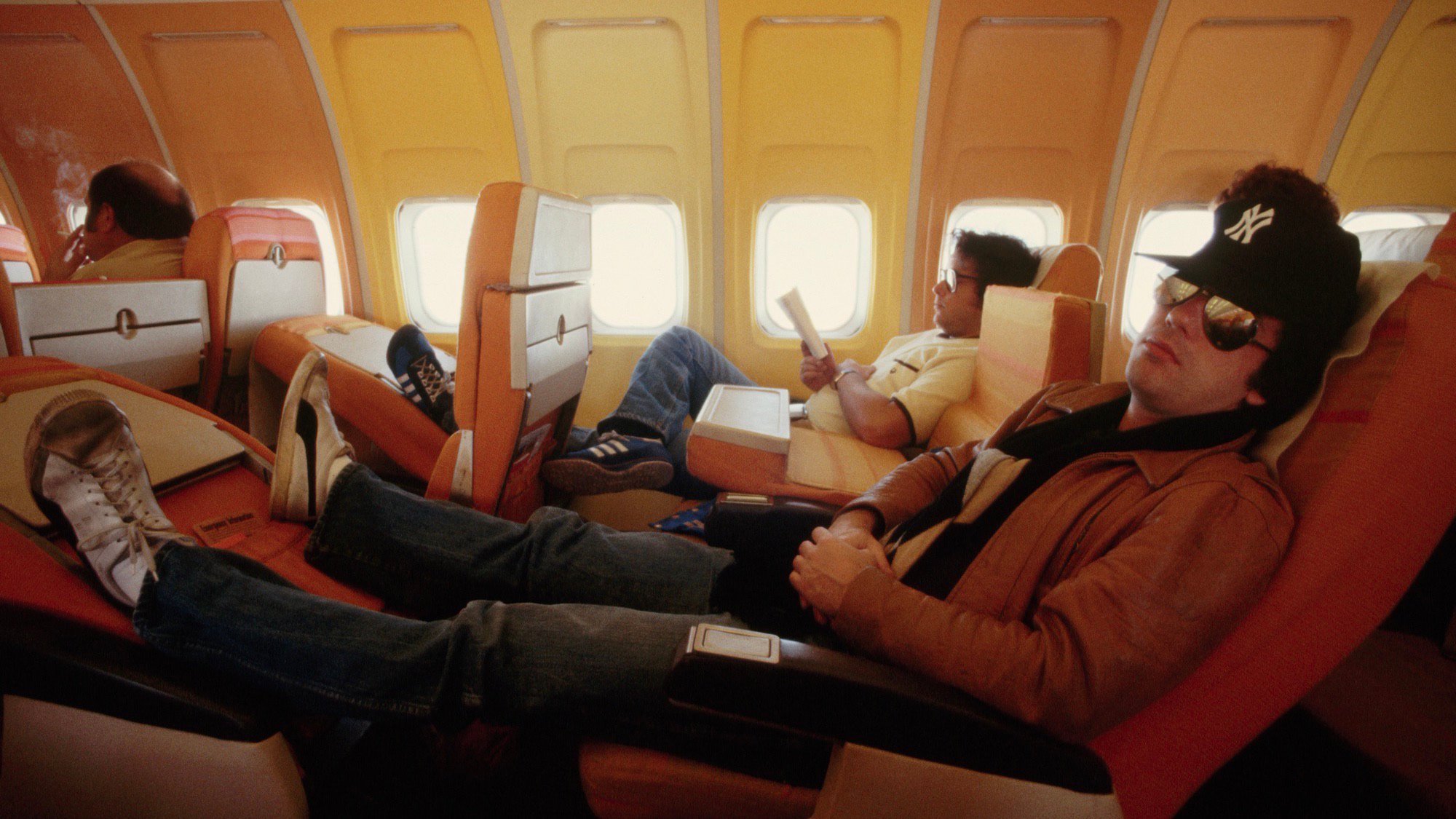 Billy Joel in the 1970s wearing a New York Yankees baseball cap, aviator sunglasses, a leather jacket, jeans, and worn-out tennis shoes. He’s napping casually on an airplane with retro orange seats and warm-toned walls ranging from orange to yellow. The seat in front of him has been folded over so he can stretch up with his legs up on the folded seat. Behind him, a passenger with glasses is reading, while another passenger in front is smoking a cigarette.
