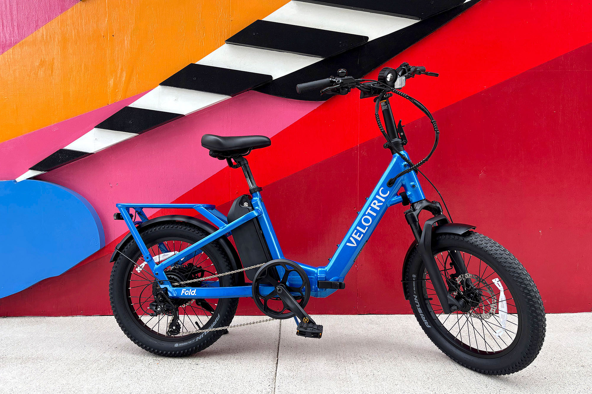 A blue Velotric Fold 1 Plus folding ebike on its kickstand in front of a colorful mural