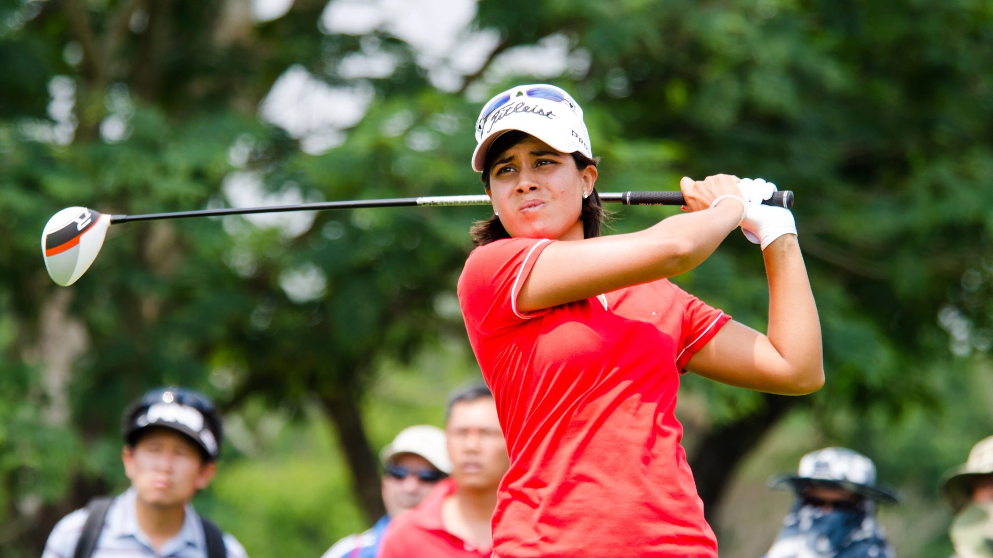 A close-up, action shot of professional female golfer Julieta Granada from Paraguay during a golf tournament. She is in mid-swing, wearing a red polo shirt, a white visor, and a white golf glove, with a determined expression on her face. In the background, a few spectators are partially visible behind a rope, with lush green trees blurred in the distance.
