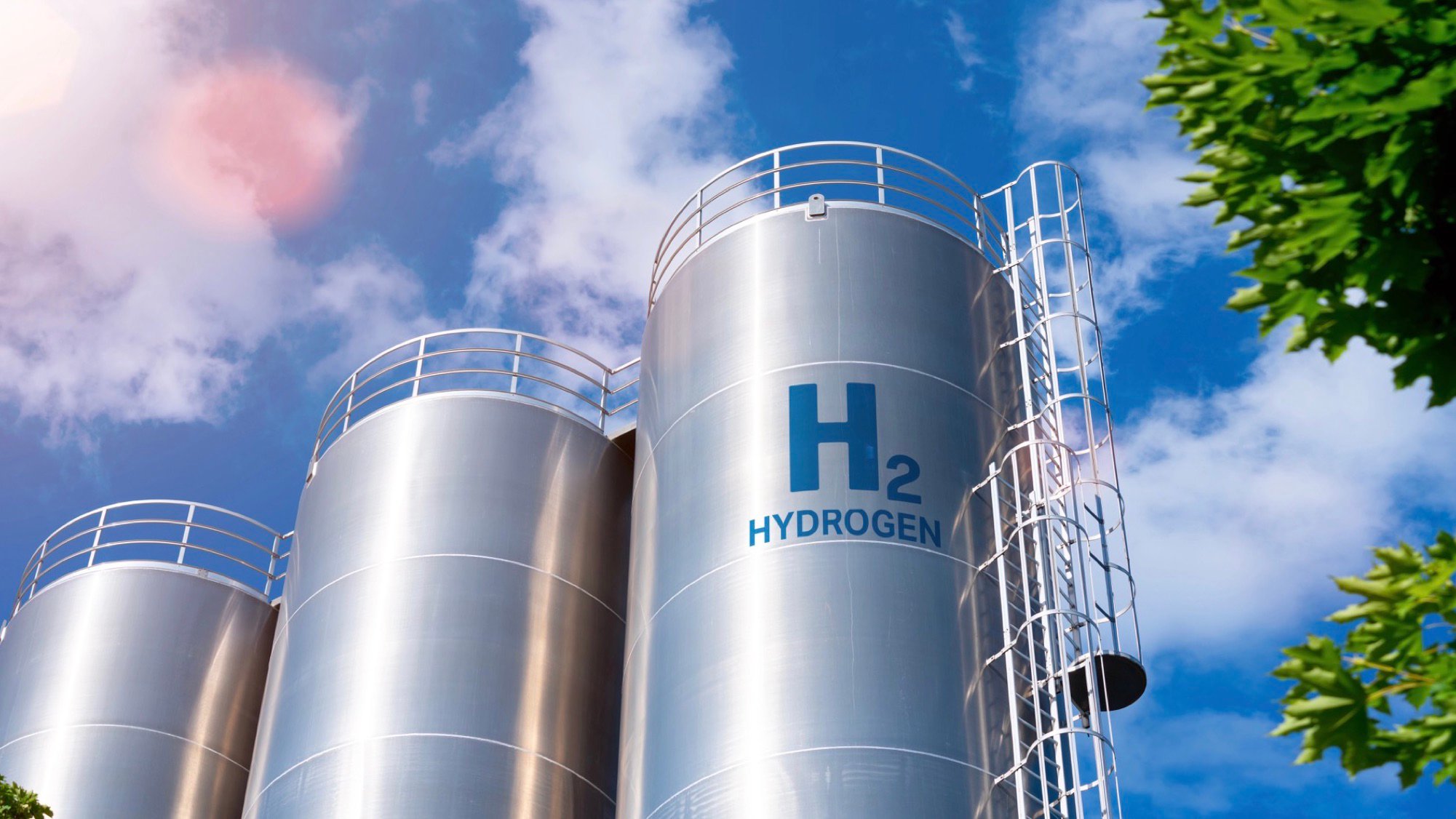 A low-angle view of three large, silver cylindrical storage tanks, with a bright blue sky and white clouds in the background. The center tank is labeled with a large "H2" and the word "HYDROGEN." A metal ladder and railing system is attached to the side of the tank. The corner of a tree with green leaves is visible in the foreground on the right.