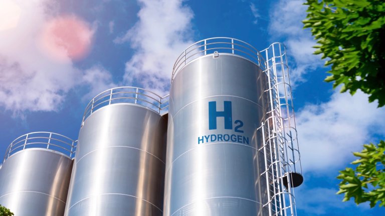 A low-angle view of three large, silver cylindrical storage tanks, with a bright blue sky and white clouds in the background. The center tank is labeled with a large "H2" and the word "HYDROGEN." A metal ladder and railing system is attached to the side of the tank. The corner of a tree with green leaves is visible in the foreground on the right.