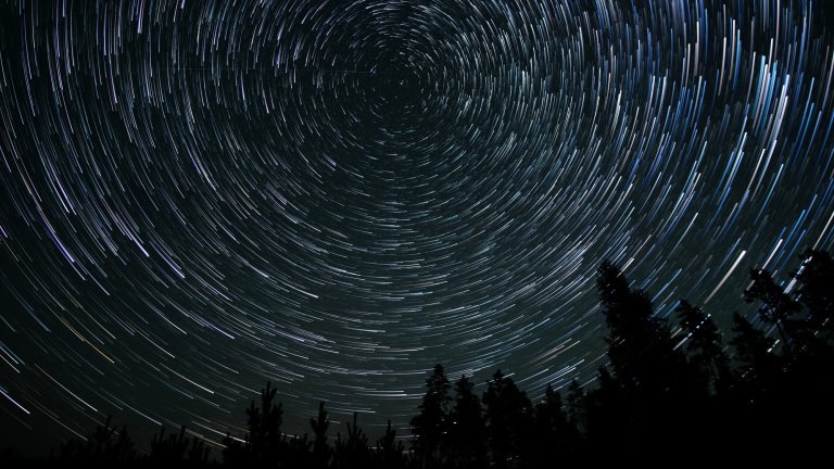 A long-exposure photograph of a night sky, showing the circular motion of stars as white streaks of light. The star trails radiate out from a central point, with some appearing thicker and brighter. In the lower part of the frame, the dark silhouette of a forest of evergreen trees is visible against the night sky.