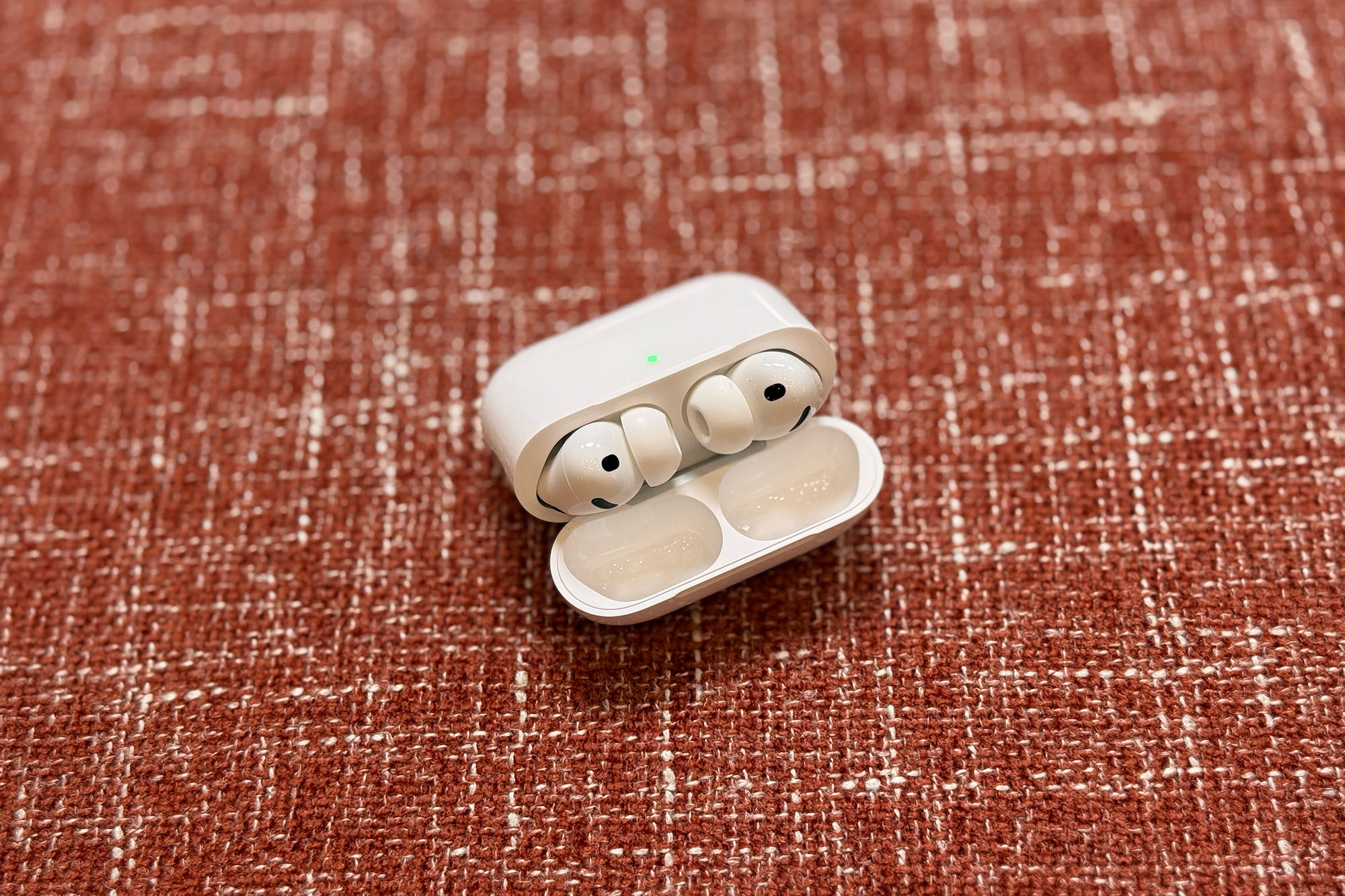 White AirPods Pro 3 earbuds in their case sitting open on red brick colored upholstery