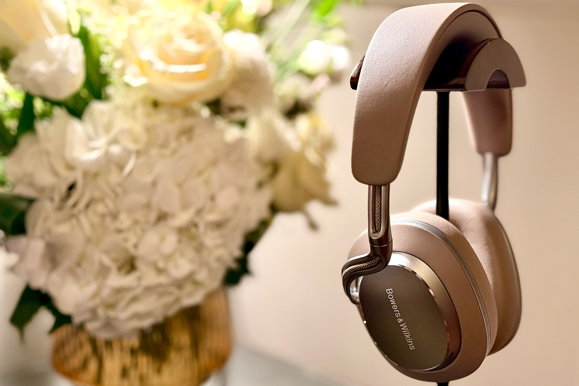 The Warm Stone Bowers & Wilkins Px8 S2 headphones on a stage in front of a floral arrangement in a New York hotel suite.